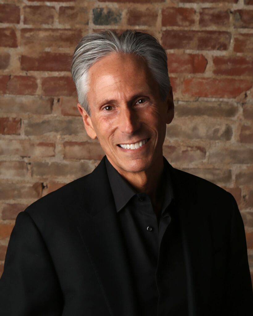 A man in a black suit smiling in front of a brick wall.