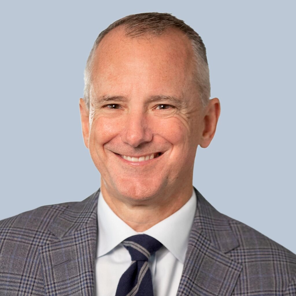 A man with short gray hair wearing a suit and tie smiles against a light gray background.