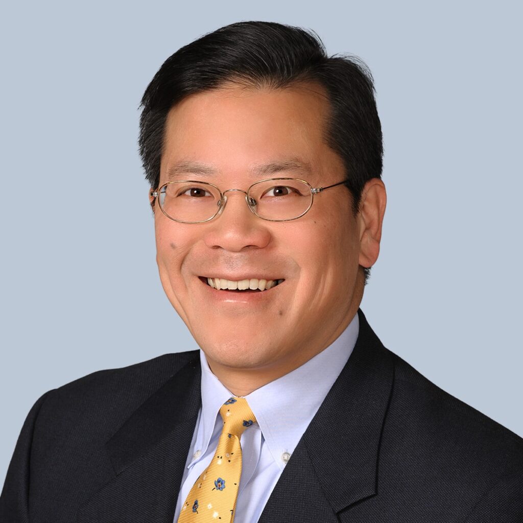 Man with short black hair and glasses wearing a dark suit, light blue shirt, and yellow tie smiles against a light blue background.