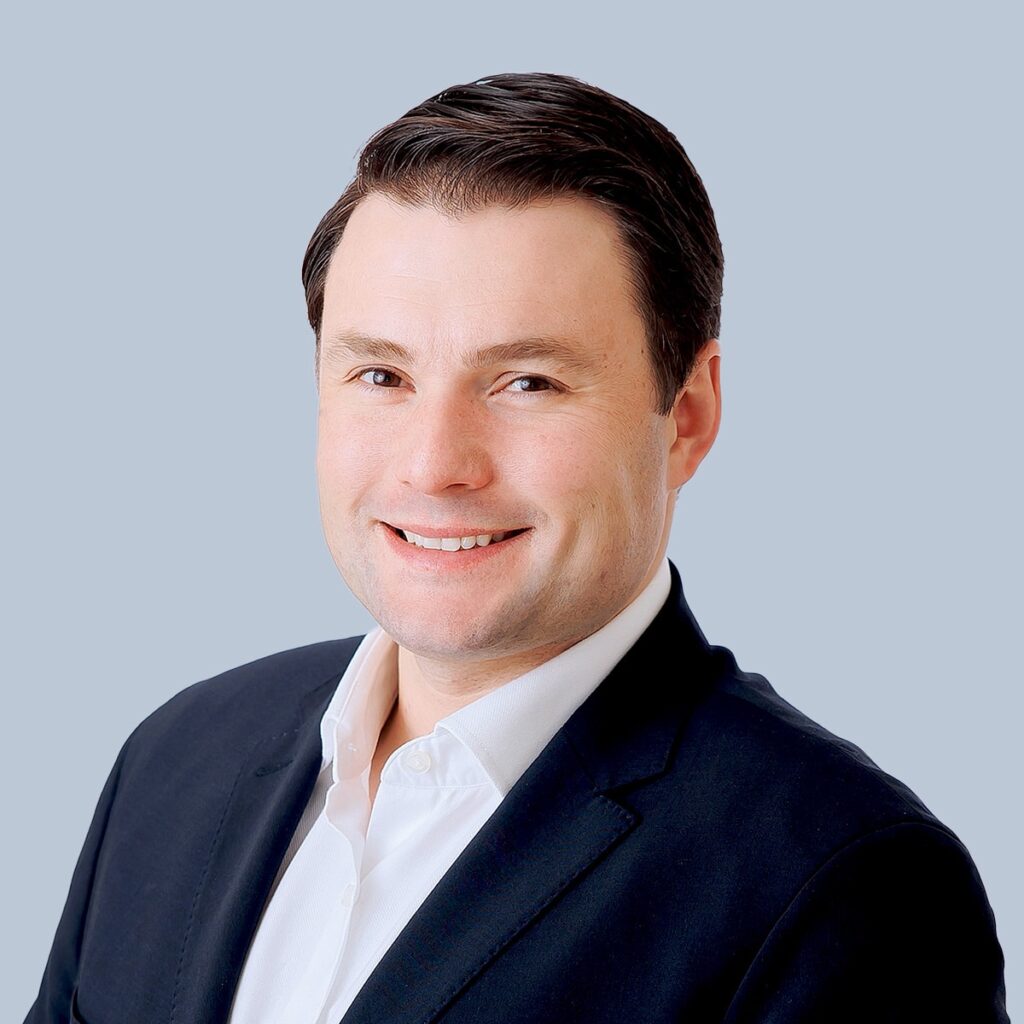 A man wearing a dark blazer and white shirt smiles at the camera against a light blue background.