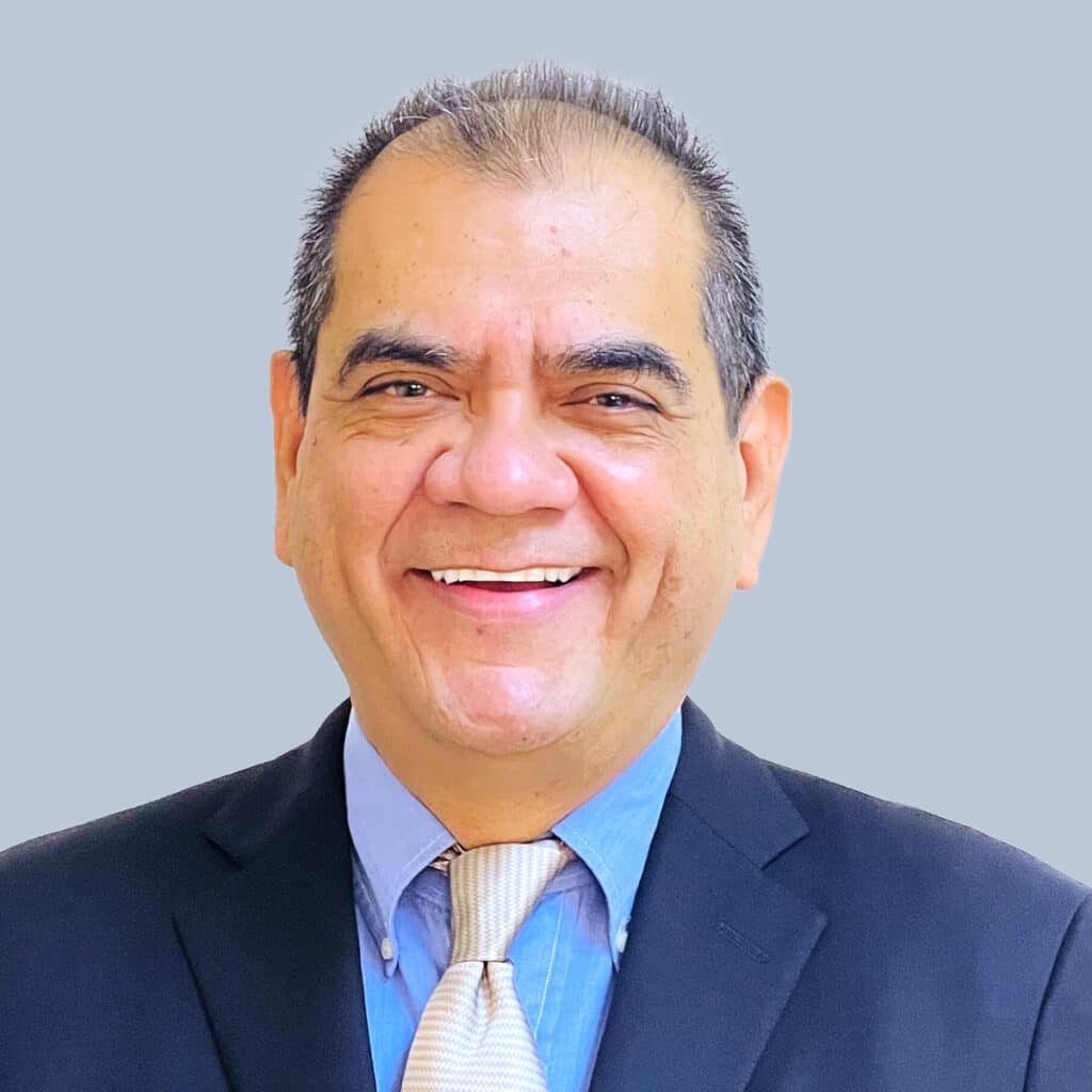 A man in a suit and tie smiles at the camera against a plain background.