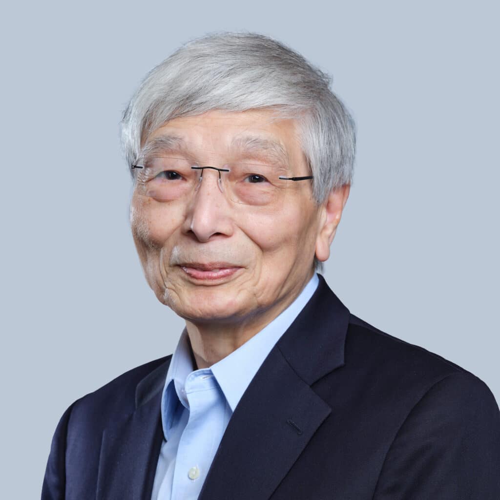 An elderly man with gray hair and glasses is wearing a dark suit jacket and light blue shirt, posing against a light gray background.