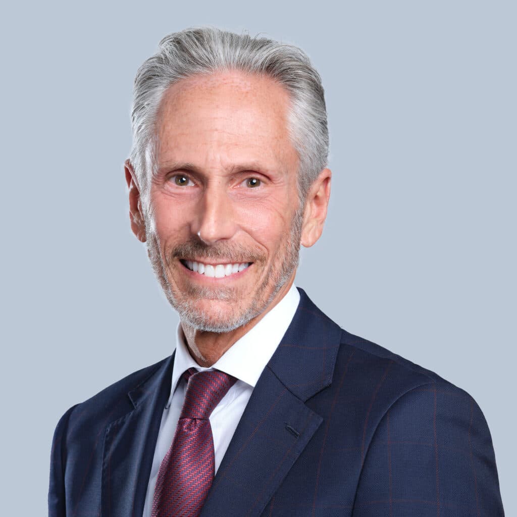 An older man with grey hair and a beard, wearing a blue suit, white shirt, and red tie, smiles at the camera against a plain background.