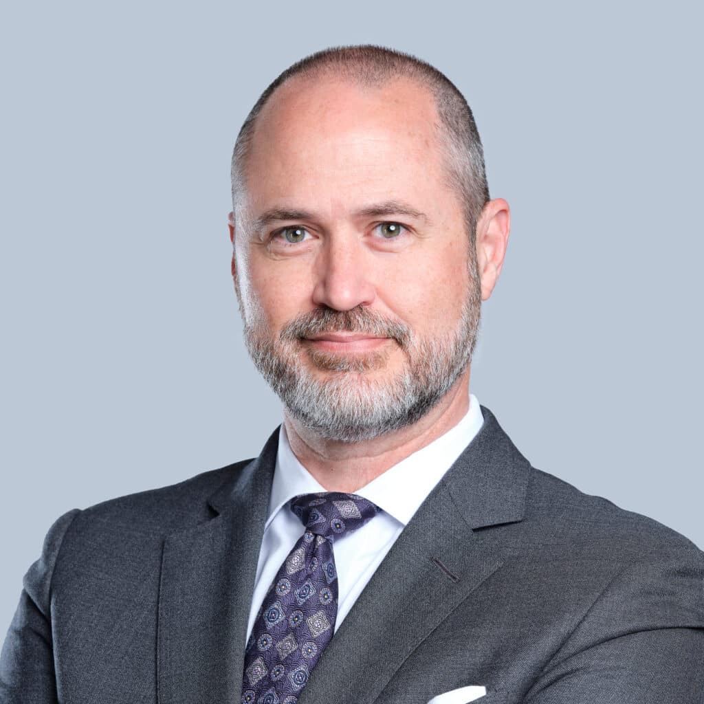 A middle-aged man with a beard and short hair, wearing a grey suit, white shirt, and patterned tie, standing against a plain grey background.