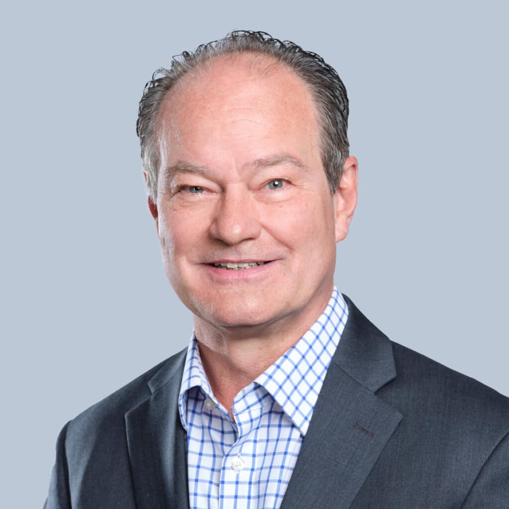 A middle-aged man with short, gray hair smiles, wearing a dark suit jacket and a blue and white checkered shirt, against a plain gray background.