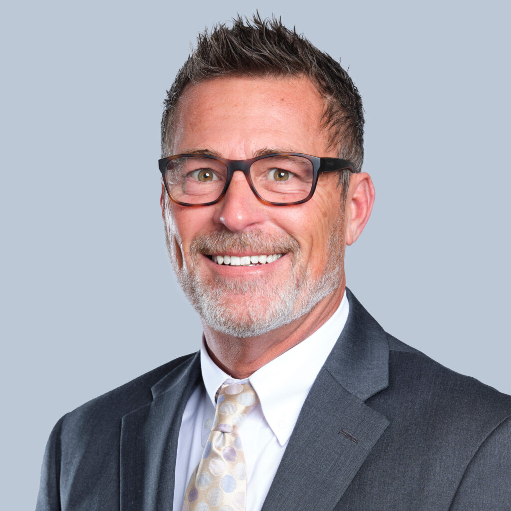 A man with short, spiked hair, glasses, and a beard, wearing a suit and a patterned tie, smiles at the camera against a plain background.