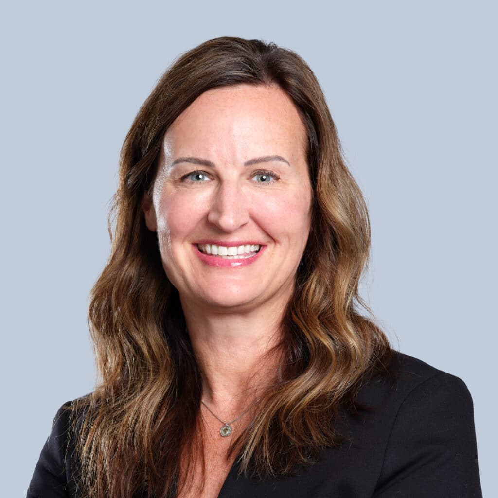 A woman with long brown hair, wearing a black blazer, smiles in front of a plain light blue background.