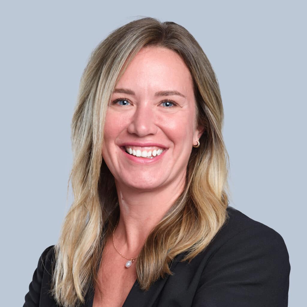 A woman with long, blonde hair wearing a black blazer smiles against a plain blue-gray background.