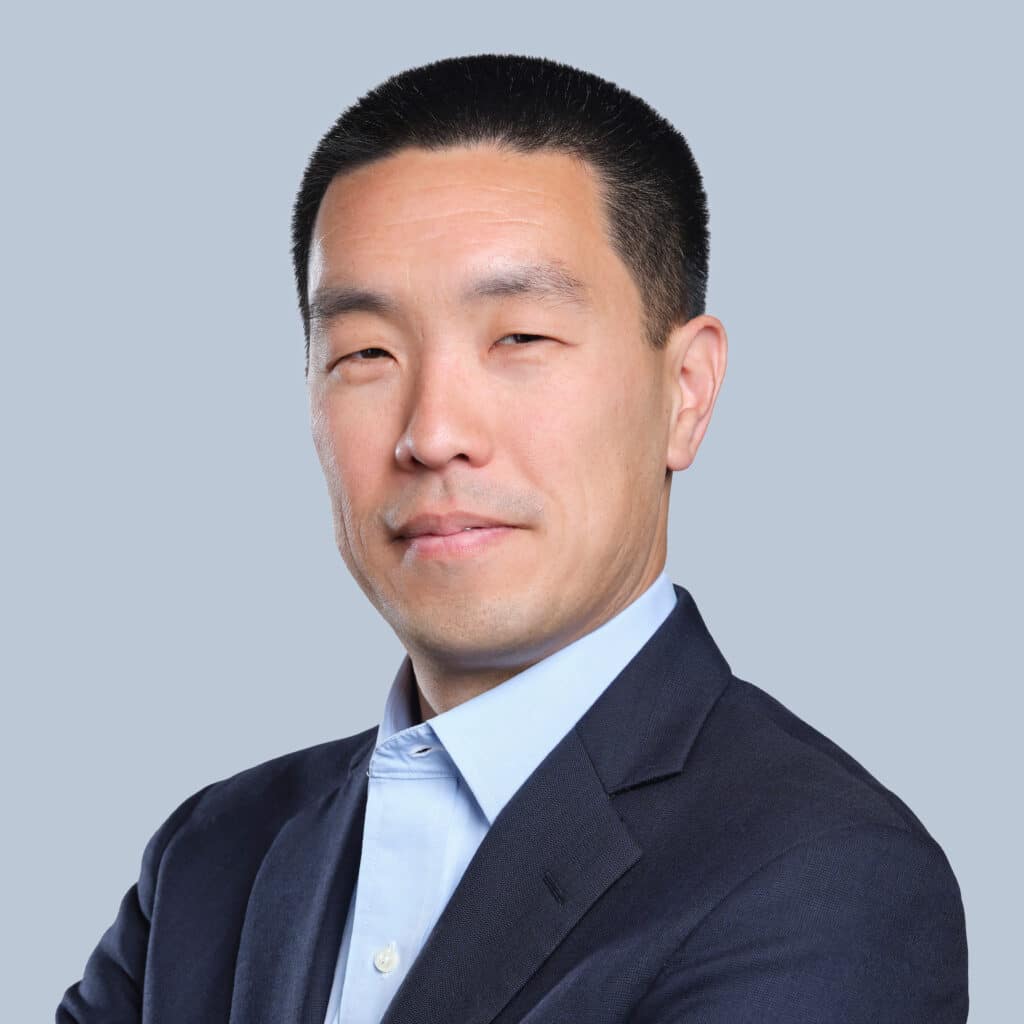 A man in a dark suit and light blue shirt stands against a plain grey background with his arms crossed.