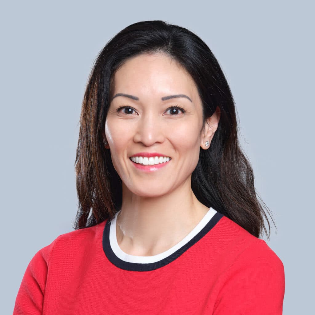 Woman with long dark hair wearing a red top, smiling against a gray background.