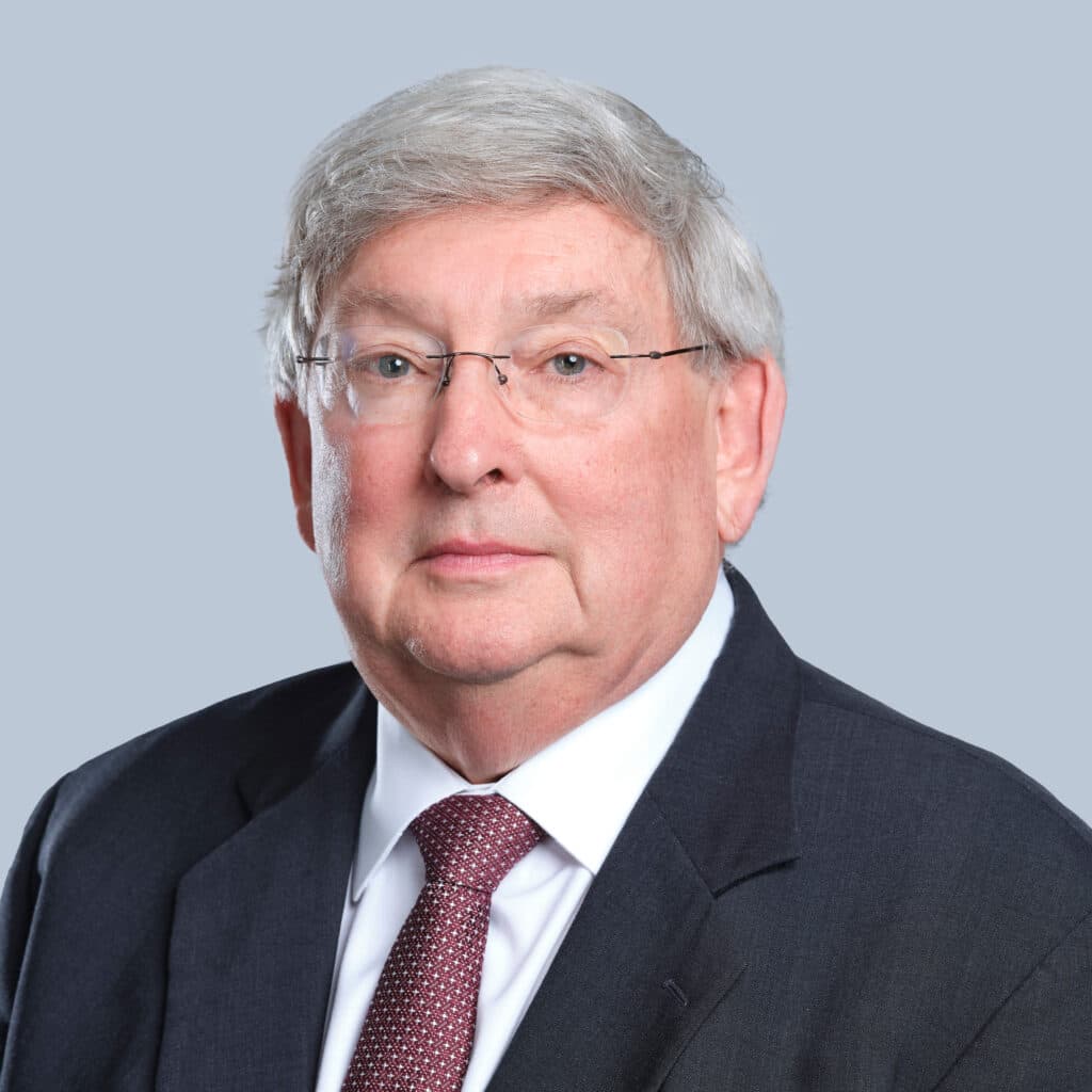 An older man with gray hair and glasses, wearing a dark suit, white shirt, and maroon tie, against a plain gray background.