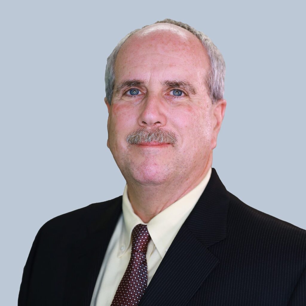 A man with short gray hair, mustache, and wearing a dark suit with a light shirt and patterned tie, poses in front of a plain background.