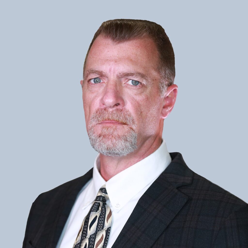 A man with short, brown hair and a goatee is seen wearing a dark suit jacket, white dress shirt, and a patterned tie, standing against a plain grey background.