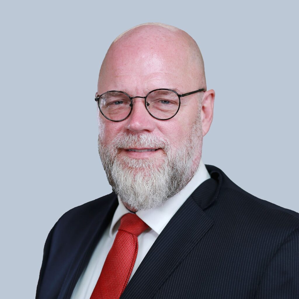 A bald man with a white beard and glasses wearing a dark suit, white shirt, and red tie standing against a light gray background.