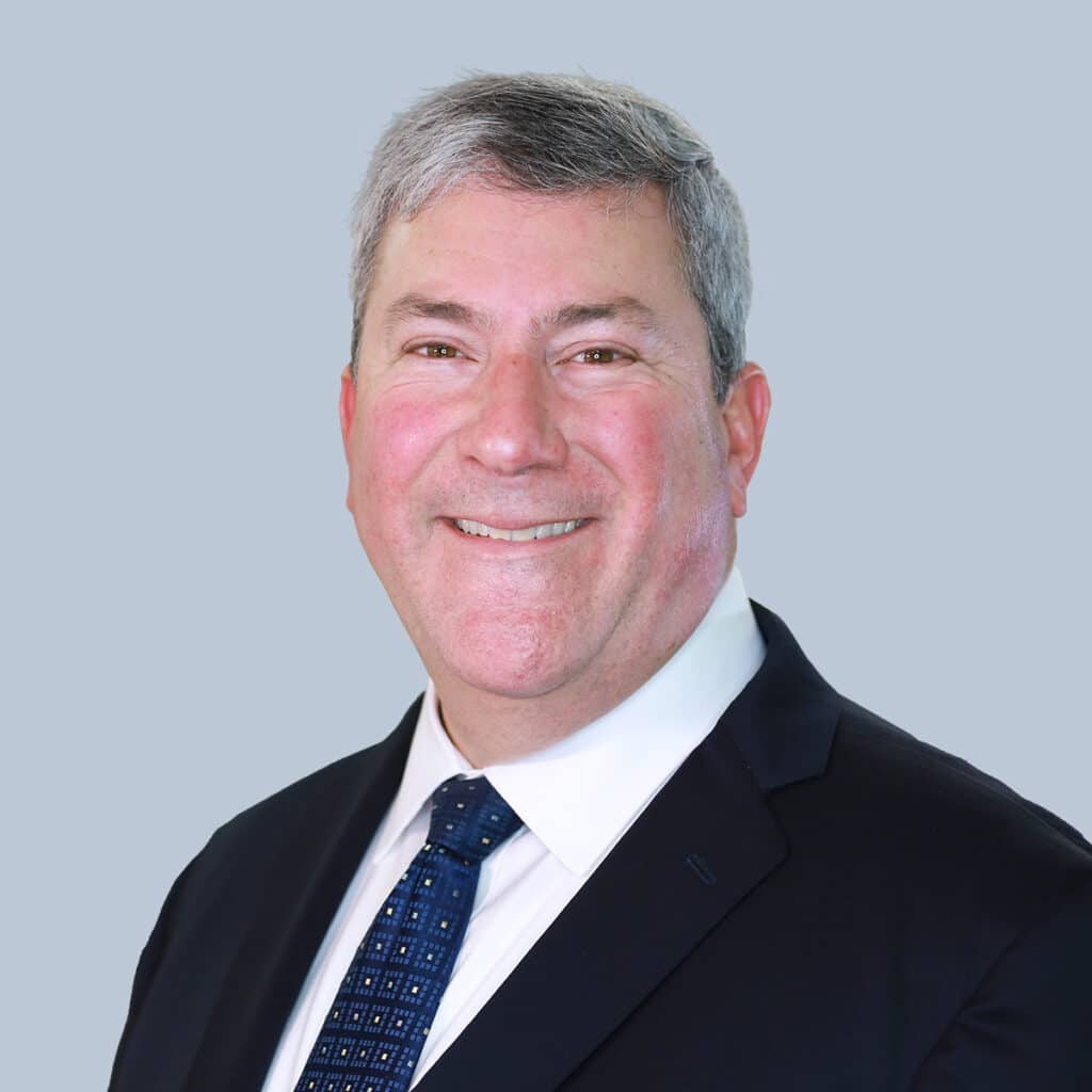 Smiling man in a suit and tie with short gray hair against a light gray background.