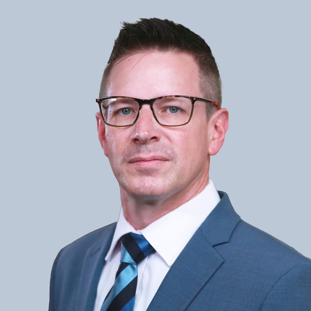 Portrait of a man wearing glasses, a blue suit, white shirt, and striped tie. He is looking directly at the camera, set against a plain gray background.