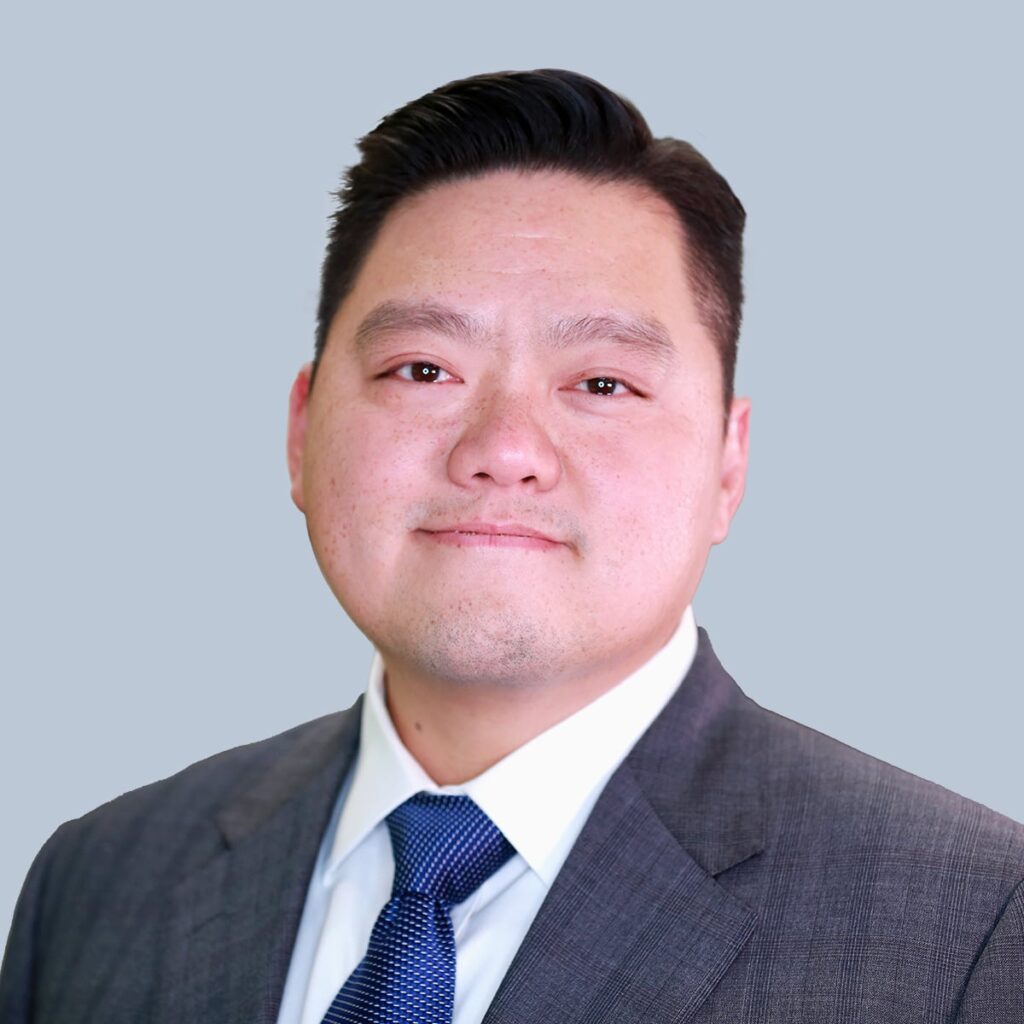 A person in a suit and tie is posing against a plain light gray background.