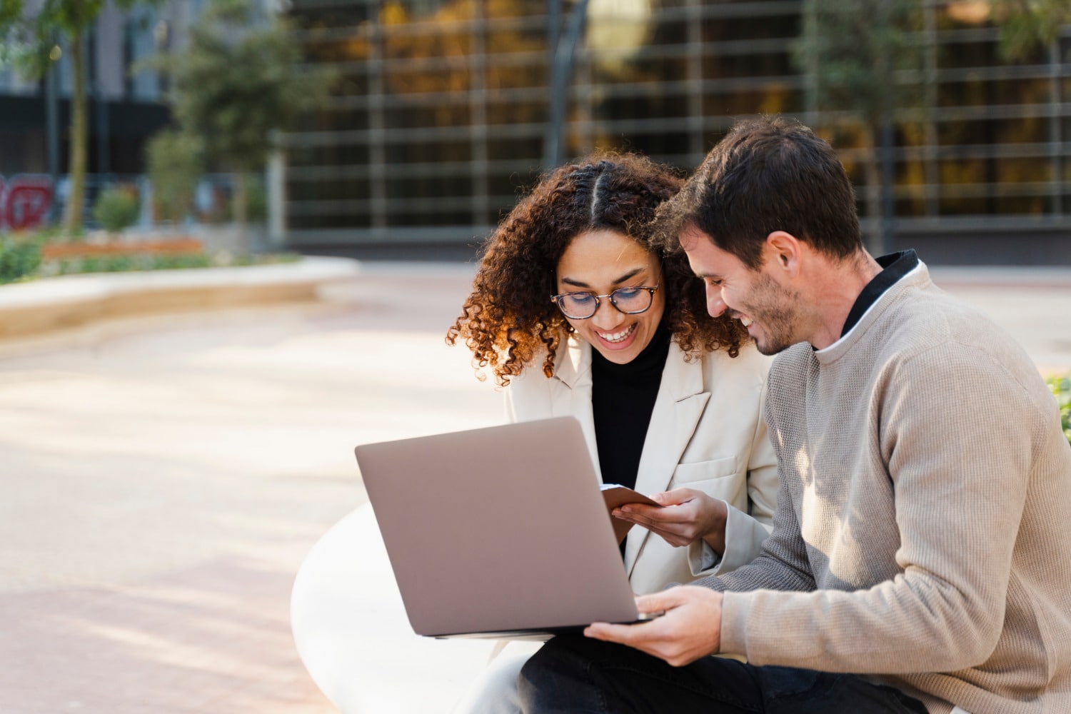 Two people sit outdoors on a bench, smiling and looking at a laptop together. One person is holding a smartphone. Trees and a glass building are in the background.