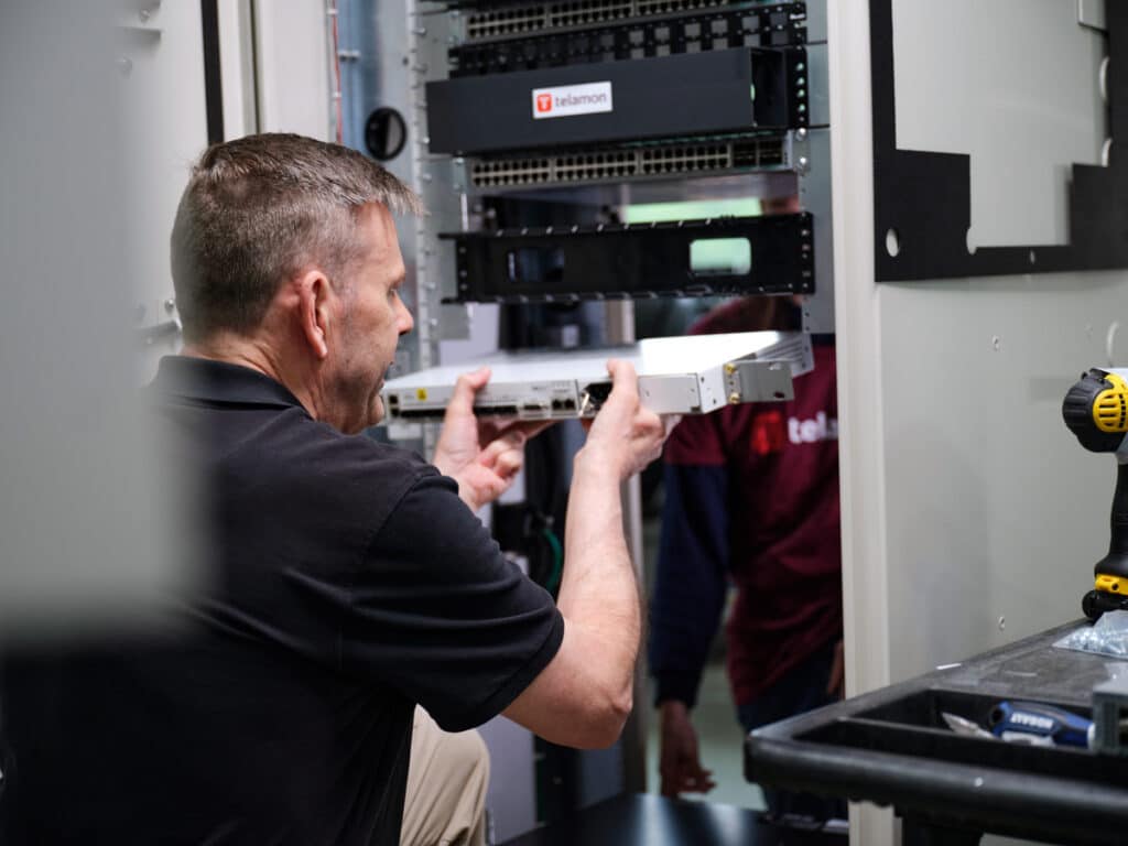 A technician installs a hardware component into a server rack. A person in the background assists with the installation.