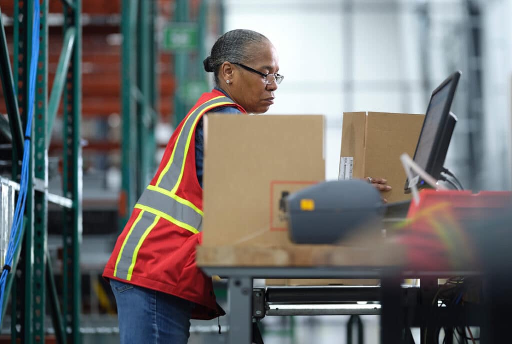 A person in a red safety vest works at a computer in a warehouse, with boxes on the table in front of them.