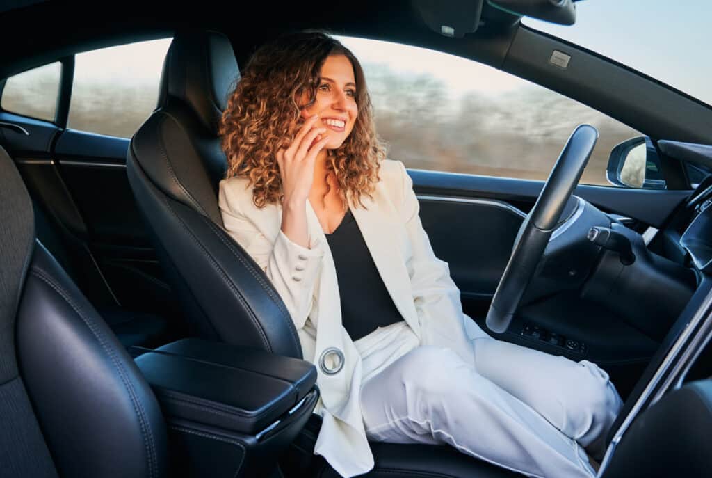 Woman with curly hair, wearing a white suit, sits in the driver's seat of a car, smiling and looking out the window while talking on the phone.