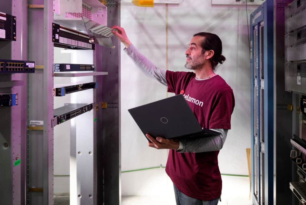 A person in a maroon shirt inspects server equipment in a data center, holding a black laptop and adjusting a piece of hardware.