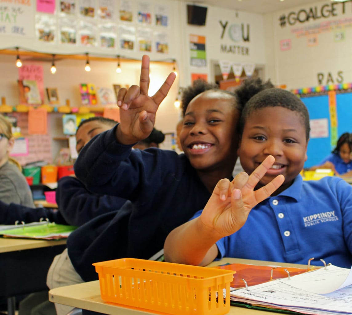 Two children seated at a classroom desk smile and make peace signs. Brightly colored books and decorations are visible in the background.