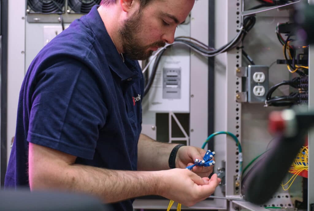 A man in a blue shirt works with wires inside an electronic equipment cabinet, focusing intently on his task.