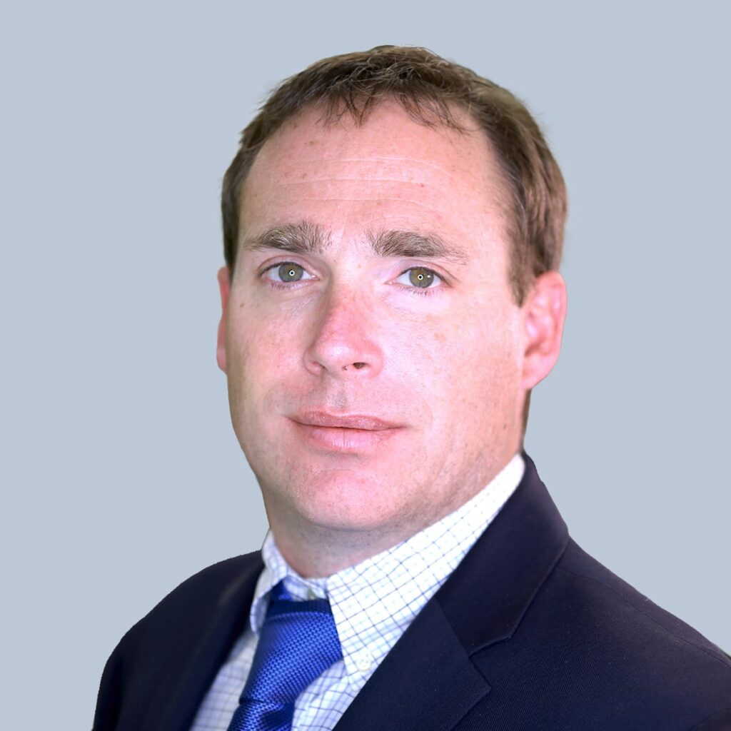 A man with short brown hair wearing a navy suit, white dress shirt, and blue tie. He is looking directly at the camera against a plain light gray background.