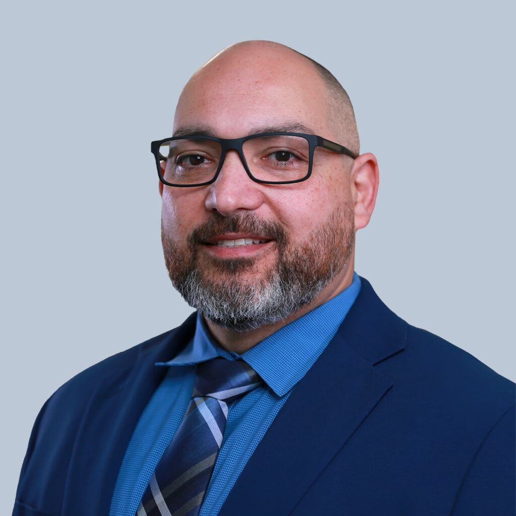 A man with a trimmed beard, glasses, and a bald head, wearing a blue suit and tie, smiles at the camera against a light blue background.