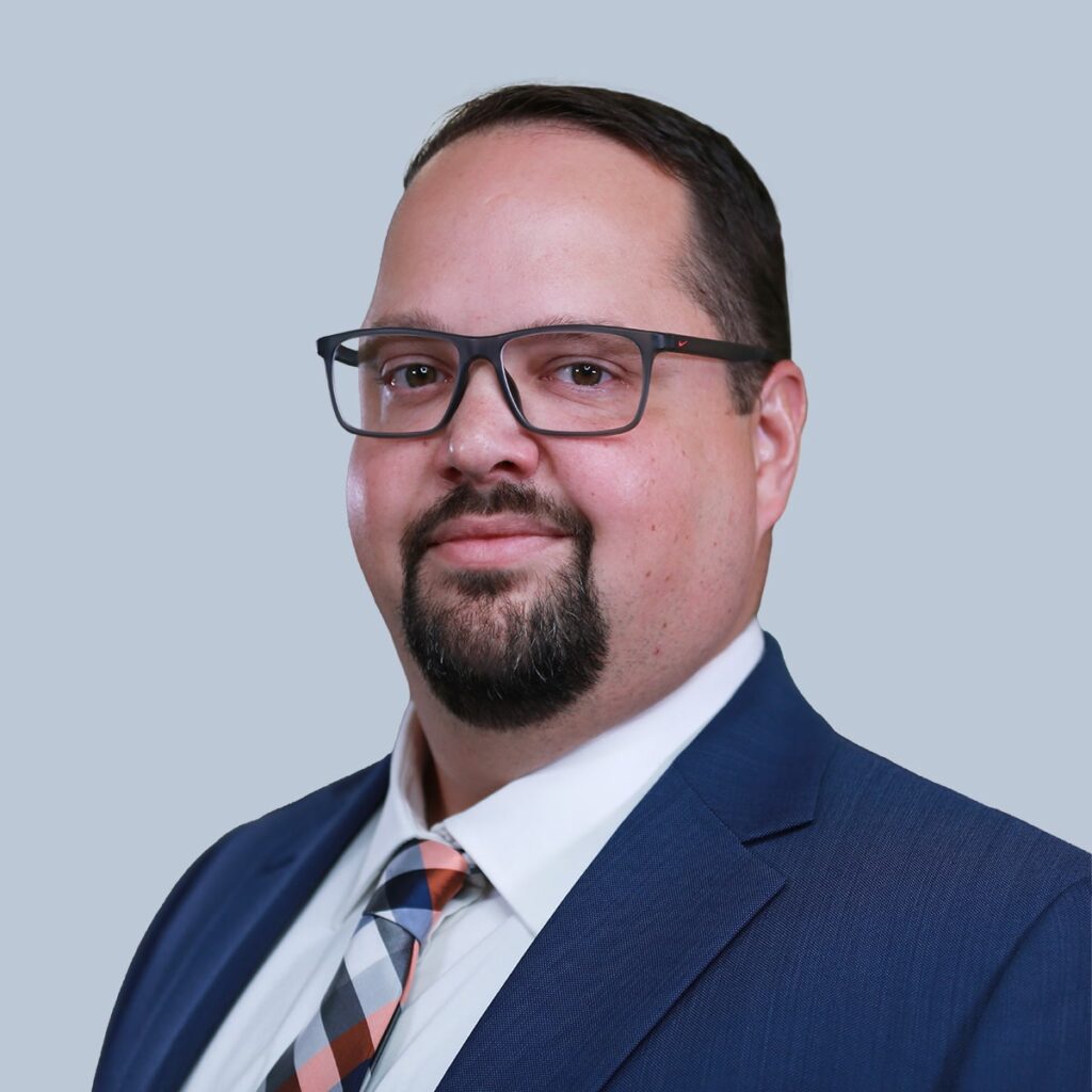 A man with glasses and a goatee wearing a dark suit, white shirt, and patterned tie, set against a light blue background.