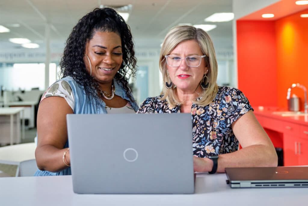 Two women in a modern office setting work together on a laptop; one woman is standing and the other is seated.