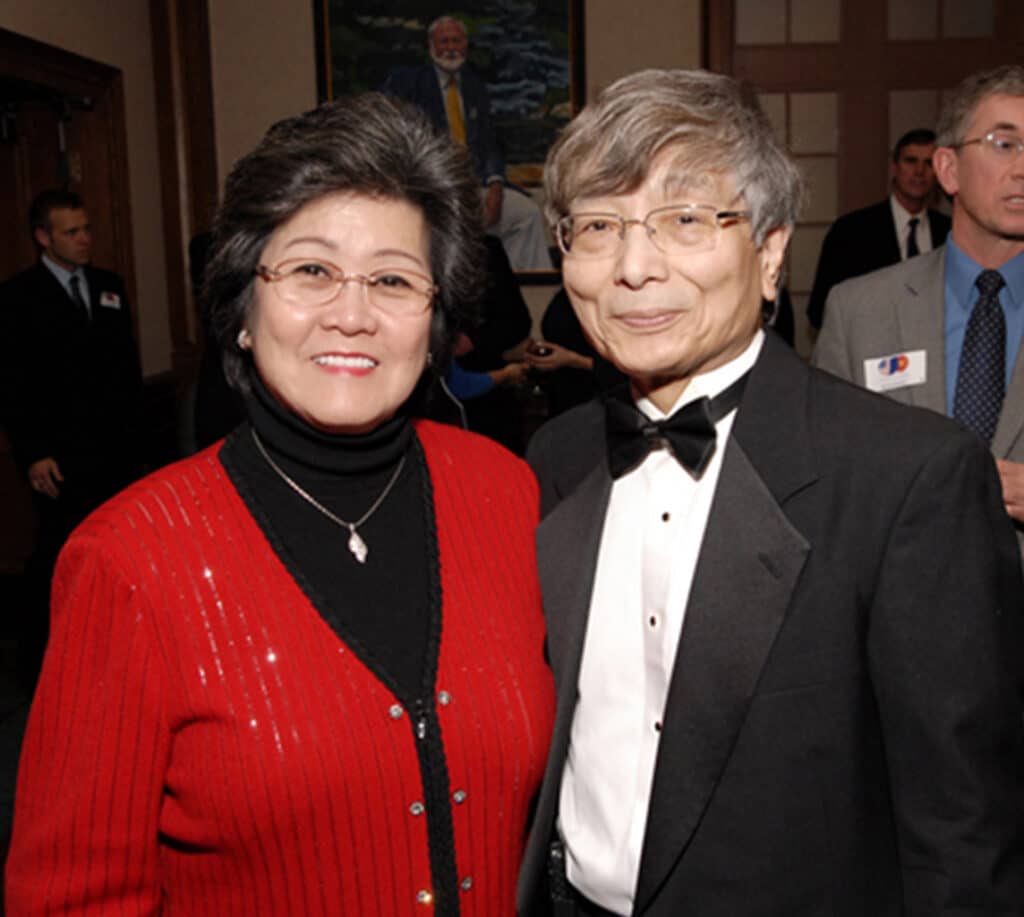 A woman in a red cardigan and man in a tuxedo pose together at an event. Other attendees are visible in the background.