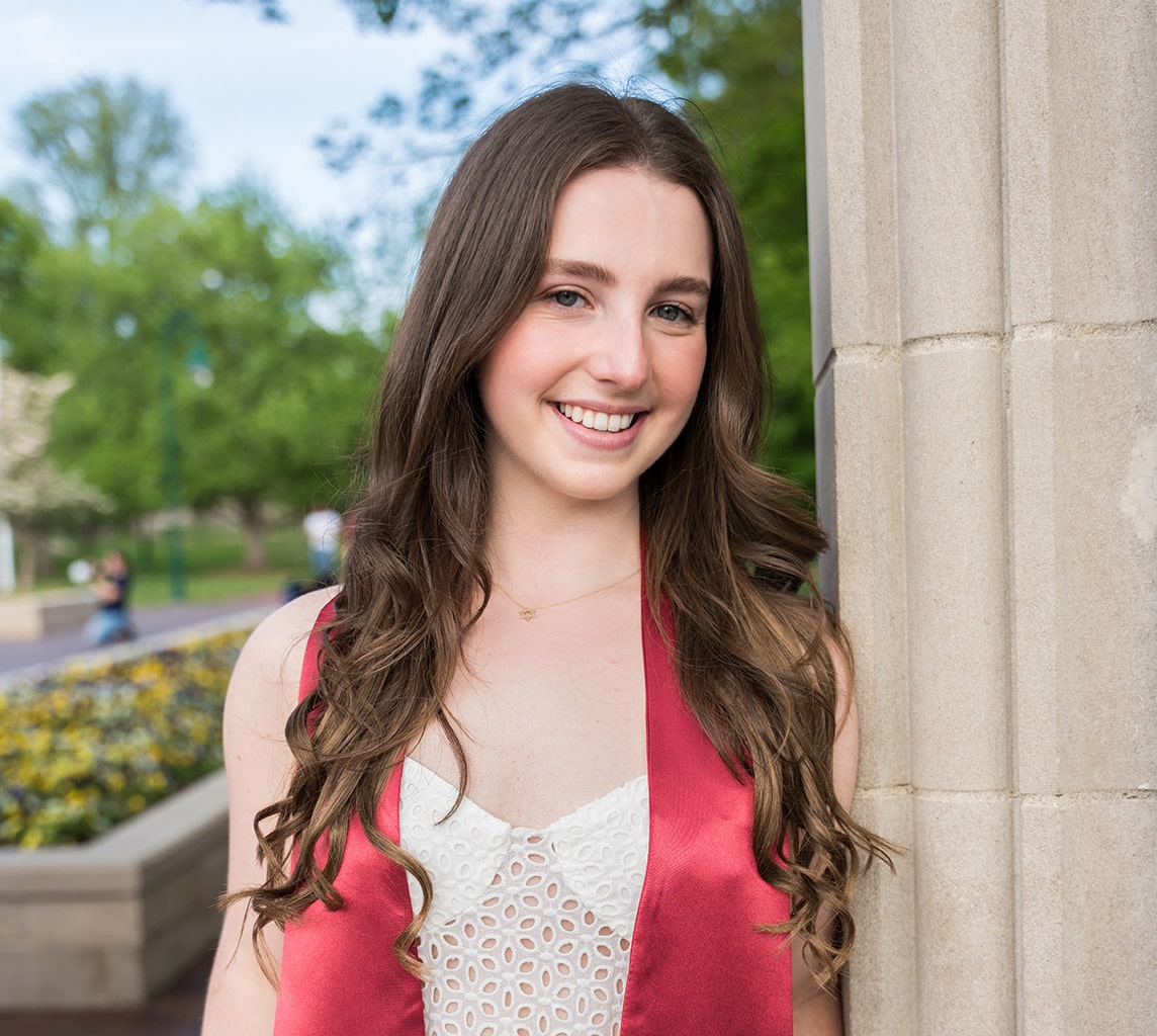 A person with long, wavy hair is standing outdoors, smiling, and wearing a red sash over a white top. There is a pillar next to them and blurry greenery in the background.