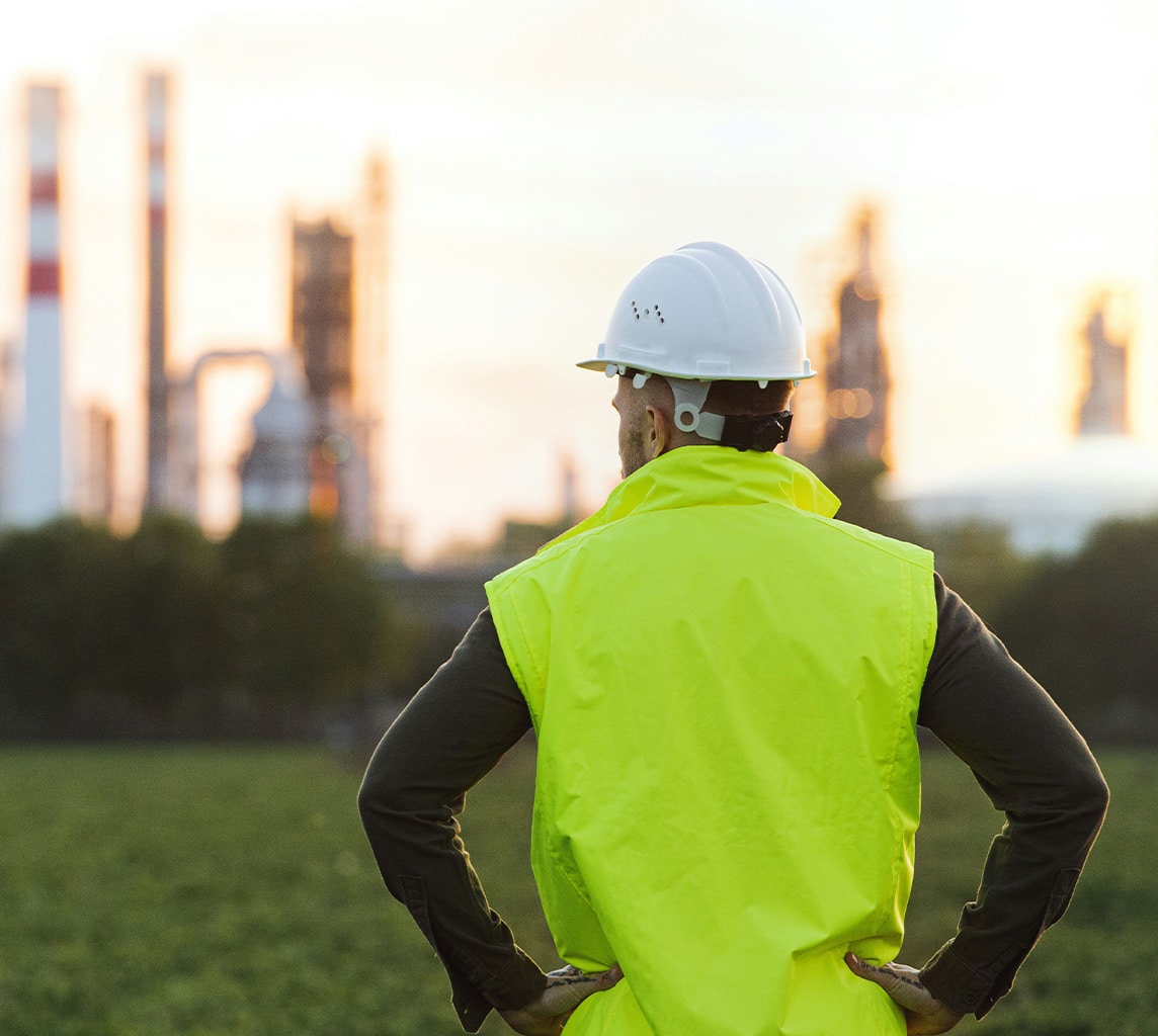 A person in a yellow safety vest and white hard hat stands with hands on hips, looking at an industrial plant in the distance.