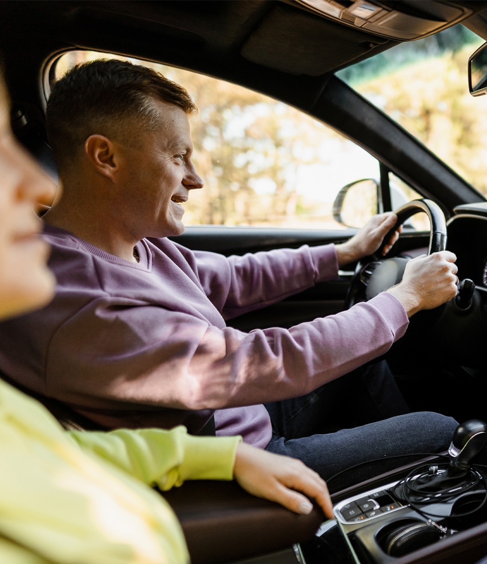 A man in a purple sweatshirt drives a car while a passenger in a light green shirt sits beside him. Sunlight filters through the window, illuminating the interior.