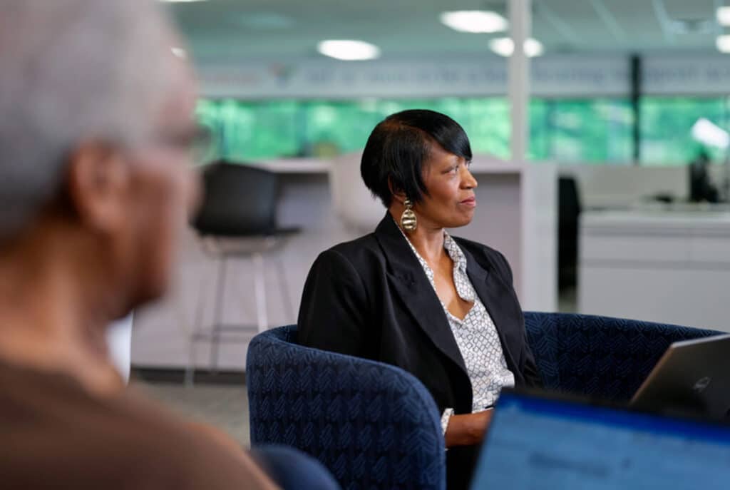 Two people are seated in an office environment. The person in the foreground is blurred, while the person in the background, dressed in business attire, is sharply focused and appears to be listening attentively.