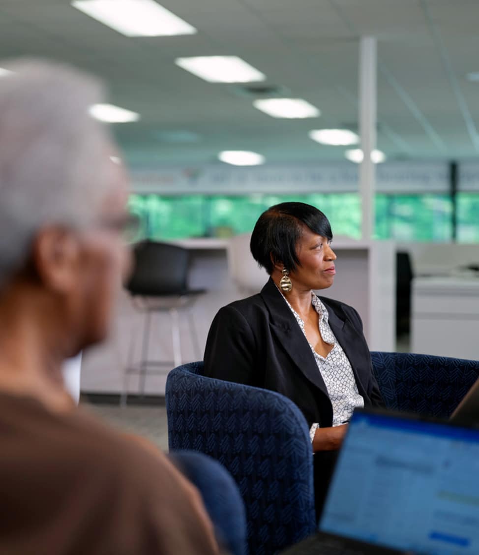 A woman in a blazer sits on a chair, facing slightly to the right, in a well-lit office space. Another person is partially visible in the foreground, working on a laptop.