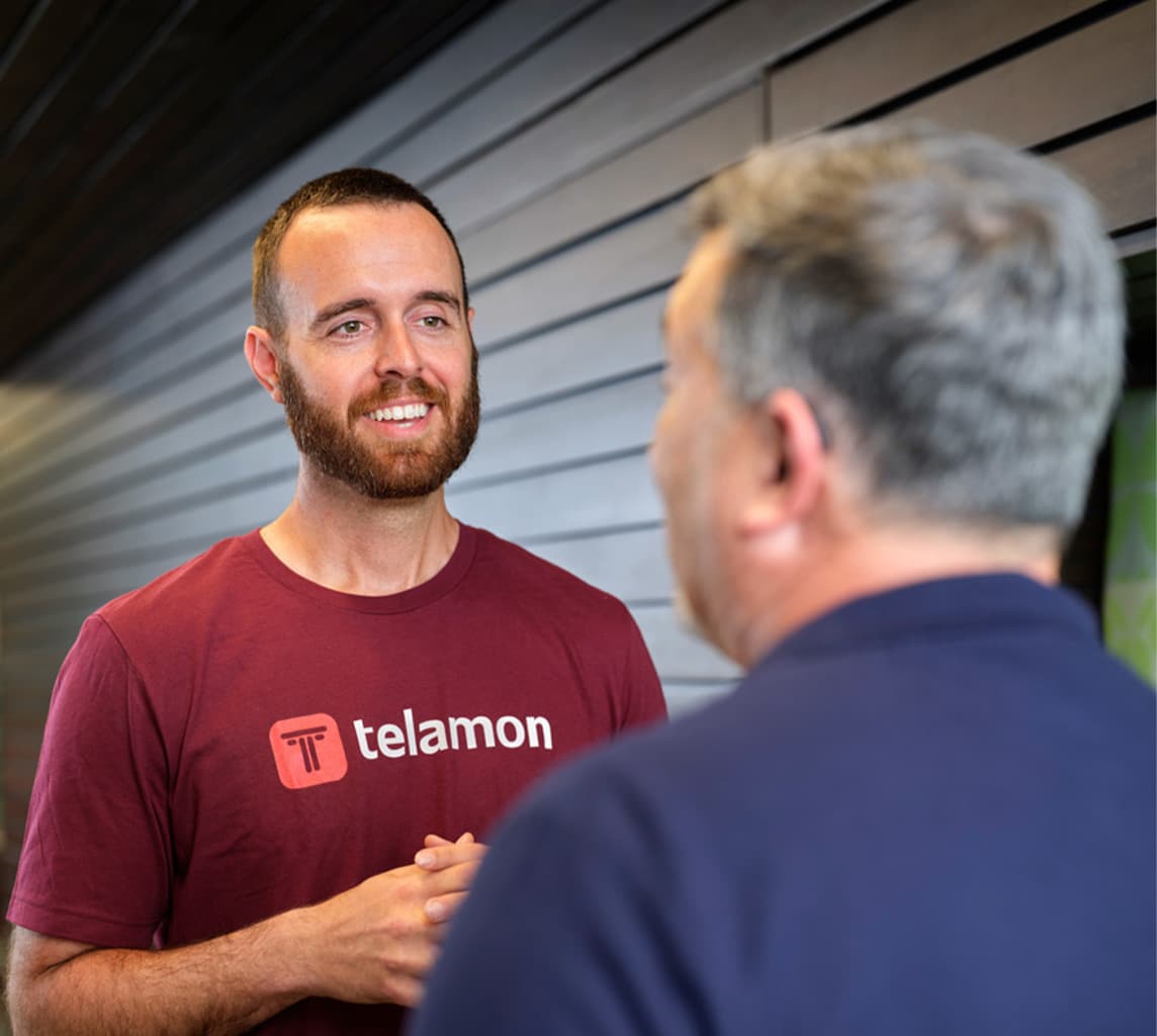 Two individuals are conversing. One of them is wearing a maroon Telamon t-shirt and smiling while speaking to the other person, who has gray hair and is seen from behind. They are indoors.