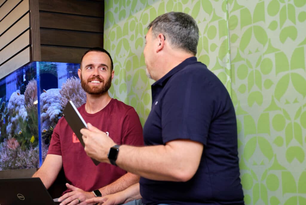 Two men sitting indoors, one in a red shirt holding a laptop and smiling, the other in a blue shirt holding a tablet, engaged in conversation in front of a fish tank and a patterned green wall.