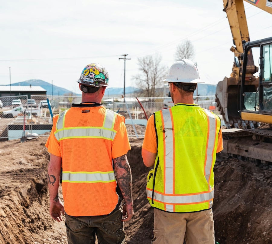 Two construction workers wearing safety vests and helmets stand on a construction site, facing an excavator and a partially dug trench.