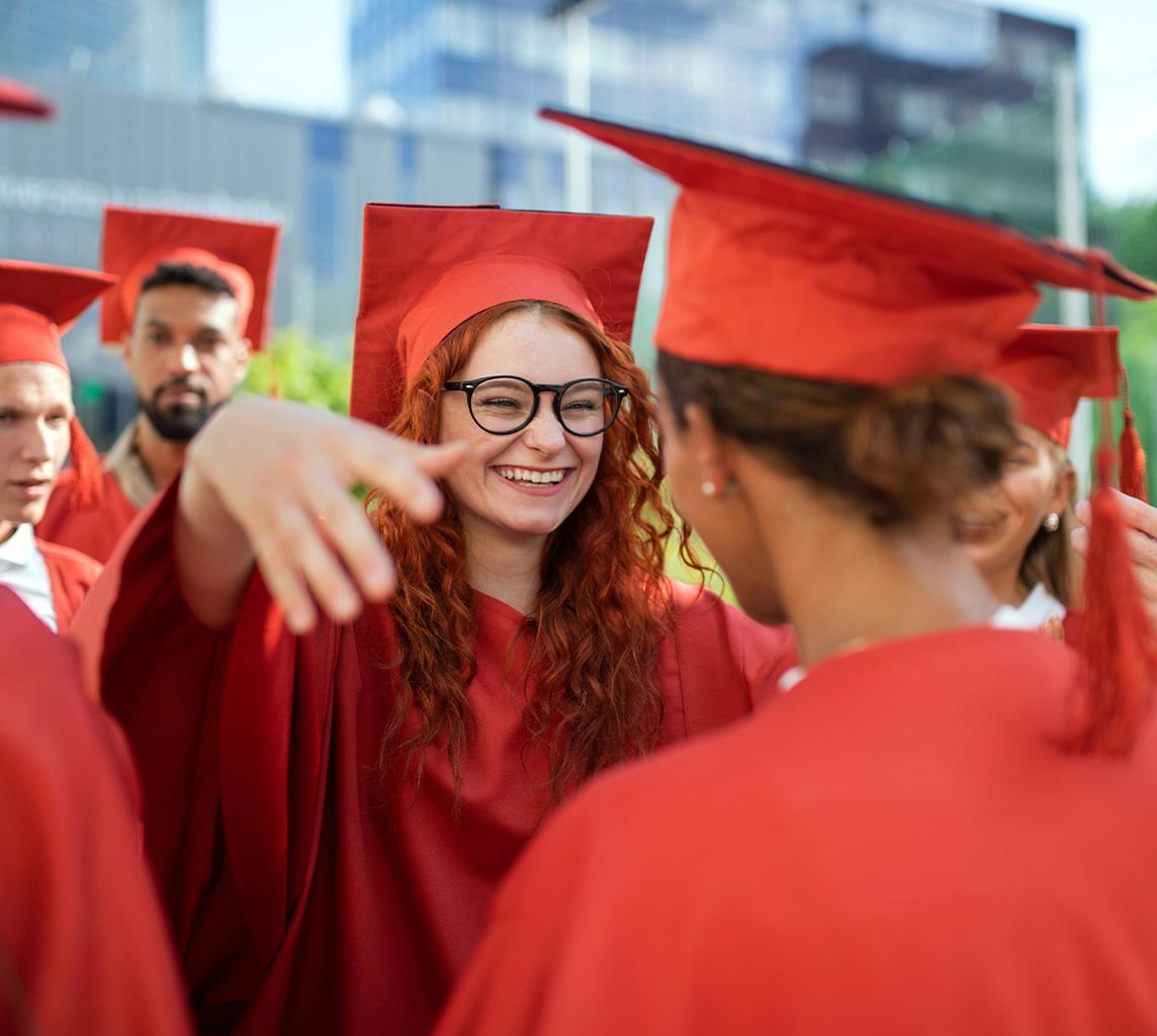 Students in red caps and gowns celebrate at an outdoor graduation ceremony. One student with glasses is smiling and has arms outstretched towards another.