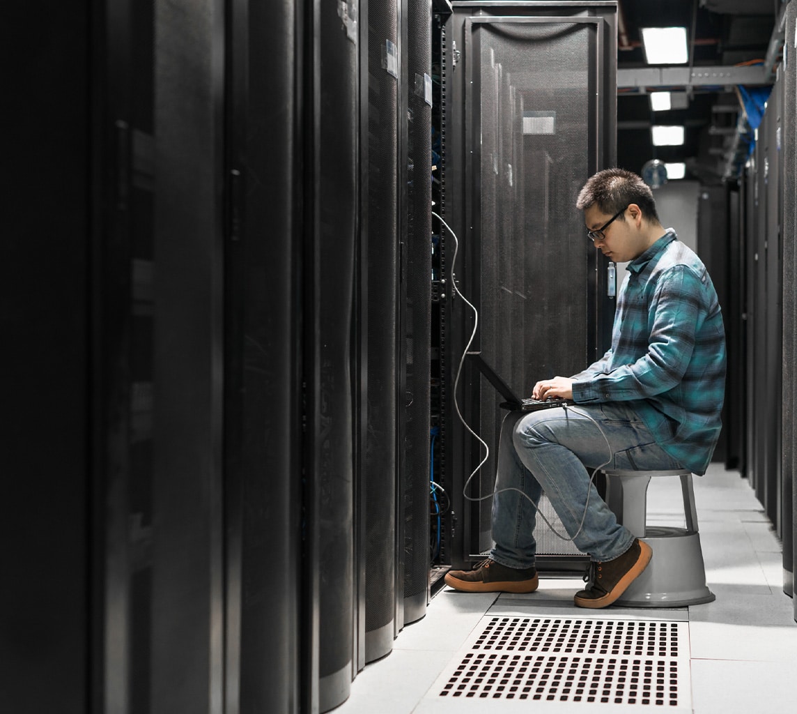 A person in a plaid shirt and jeans works on a laptop while sitting on a stool in a server room surrounded by computer servers.