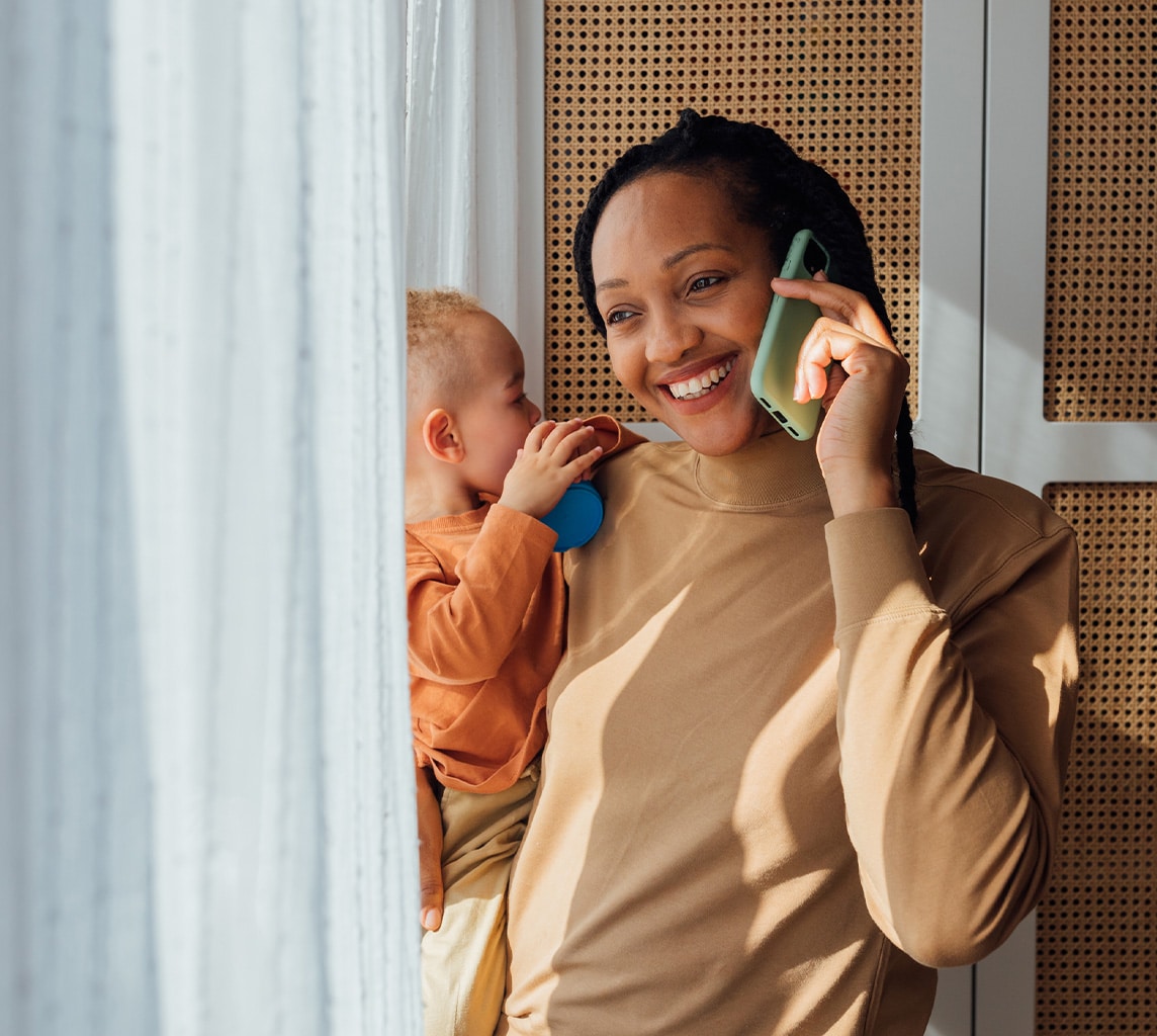 A person stands holding a baby while talking on the phone, smiling and looking outside near a window with light curtains.