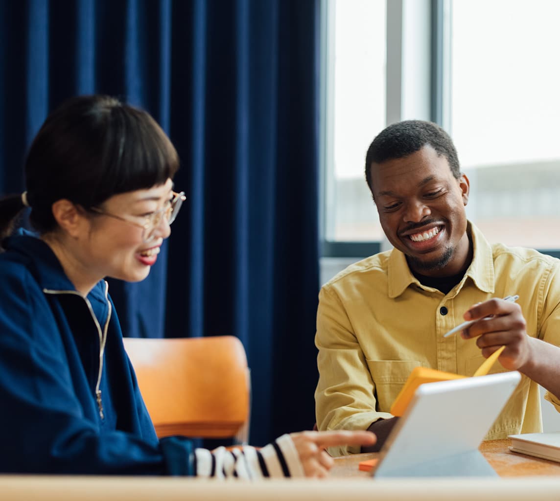 Two people sit at a table, smiling and discussing something on a tablet. One person points at the tablet screen while the other looks on, both appearing engaged and happy.