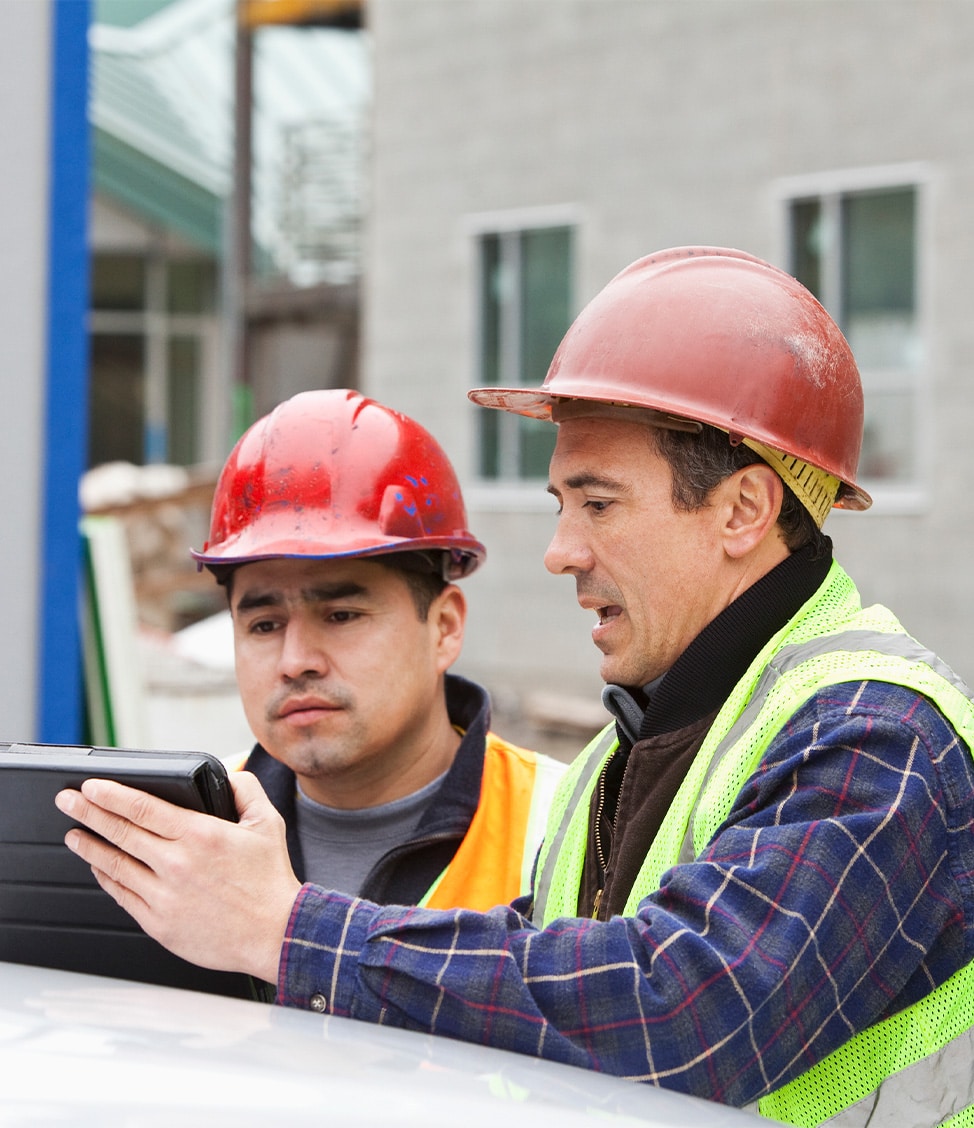 Two construction workers in hard hats and safety vests look at a tablet on a construction site.