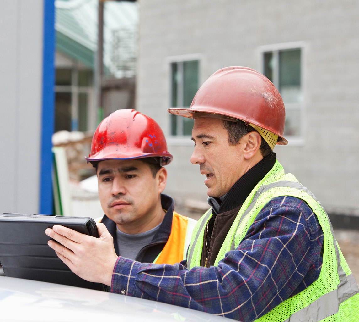 Two construction workers wearing safety vests and hard hats are looking at a tablet on a construction site. One worker is pointing at the tablet screen.