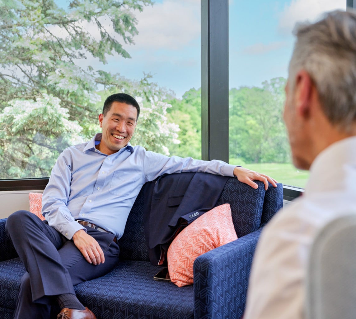 Two men are engaged in a conversation, sitting on a blue couch near large windows overlooking trees. One man is smiling, leaning back with his arm on the couch, while the other is turned towards him.