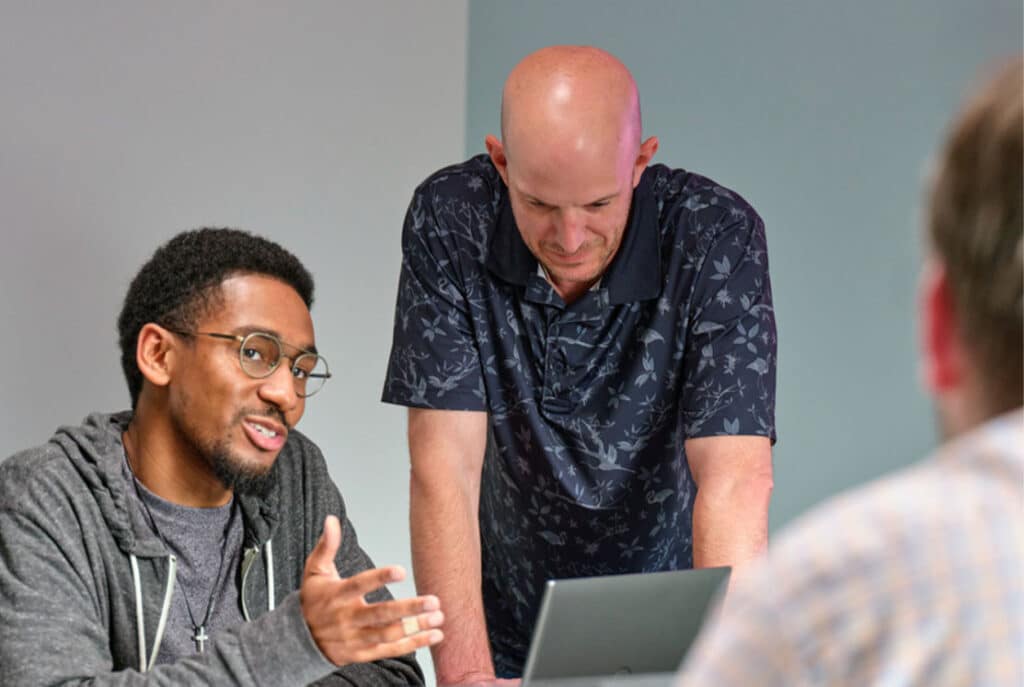 Two men are engaged in a discussion while looking at a laptop, with a third person in the foreground slightly blurred. The man on the left is speaking and gesturing, while the man on the right is leaning forward.