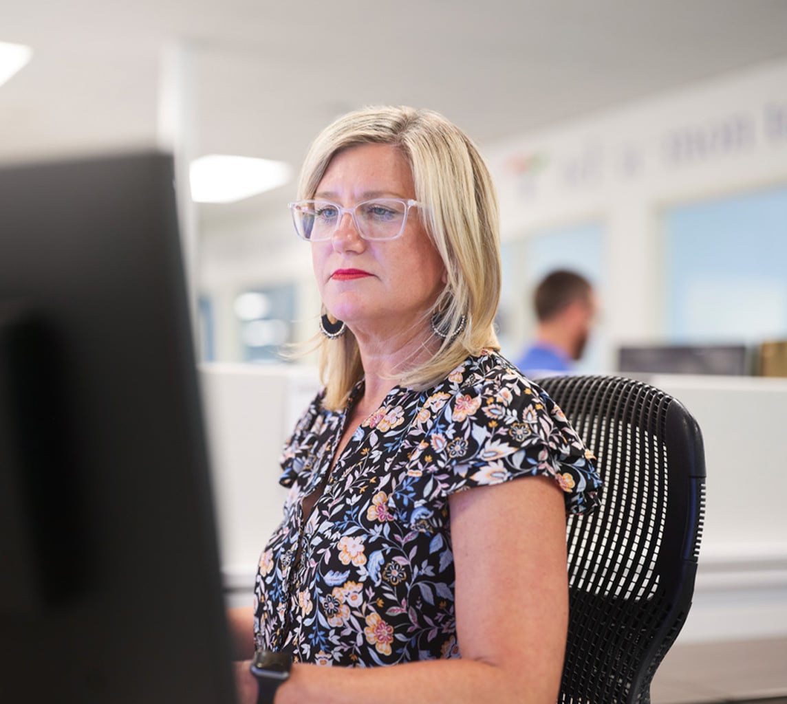 Woman with glasses and floral blouse working at a computer in an office setting. Another person blurred in the background.